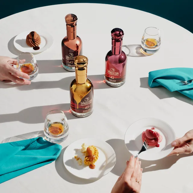 Discover the Collection Three bottles of The Singleton Gourmand Collection sit on a white tablecloth, surrounded by three plates of cakes with rocks glasses containing whisky.