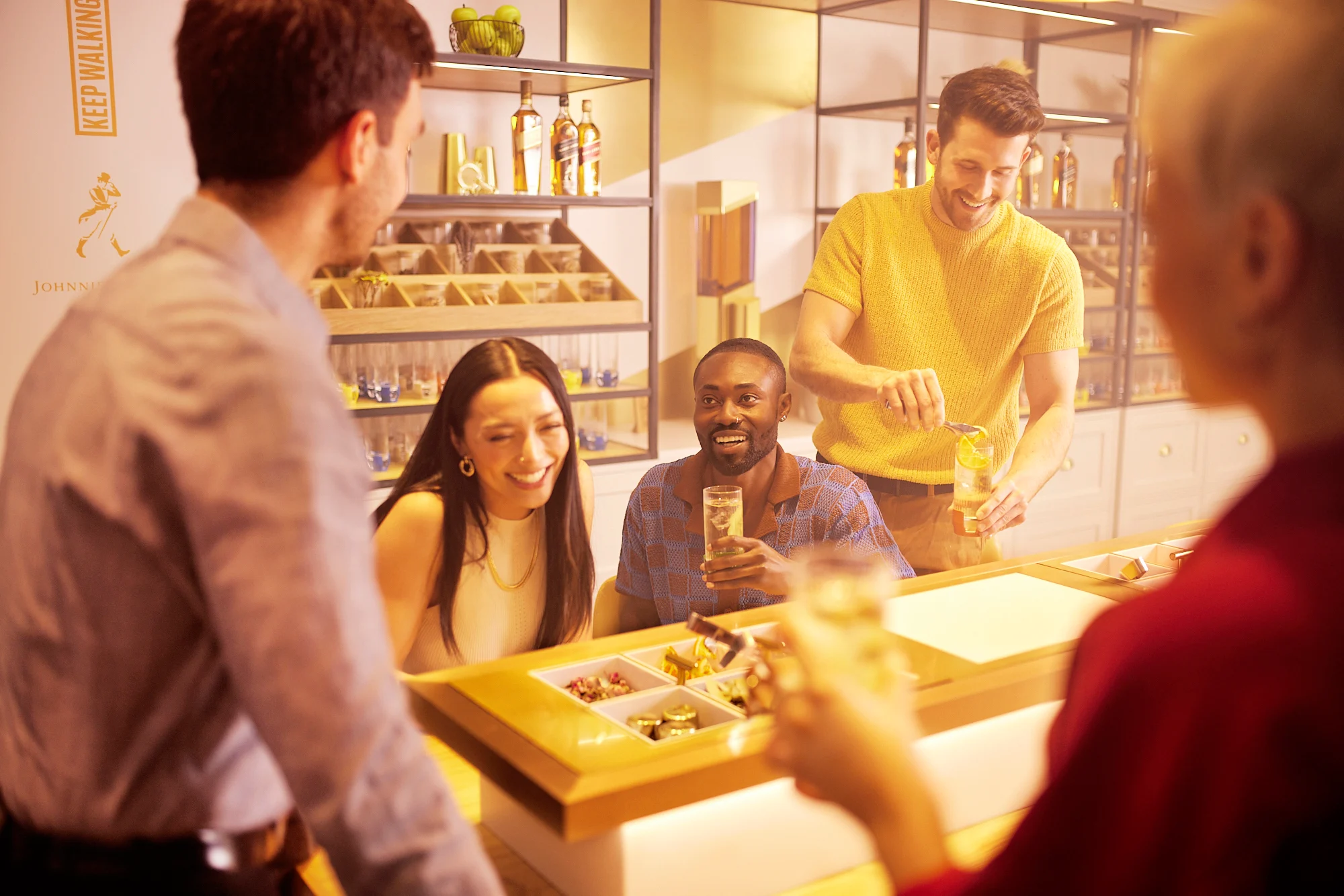 Group of friends doing the Flavour of Journey tour smiling and preparing their highball cocktails while sitting around the table with different cocktail ingredients 