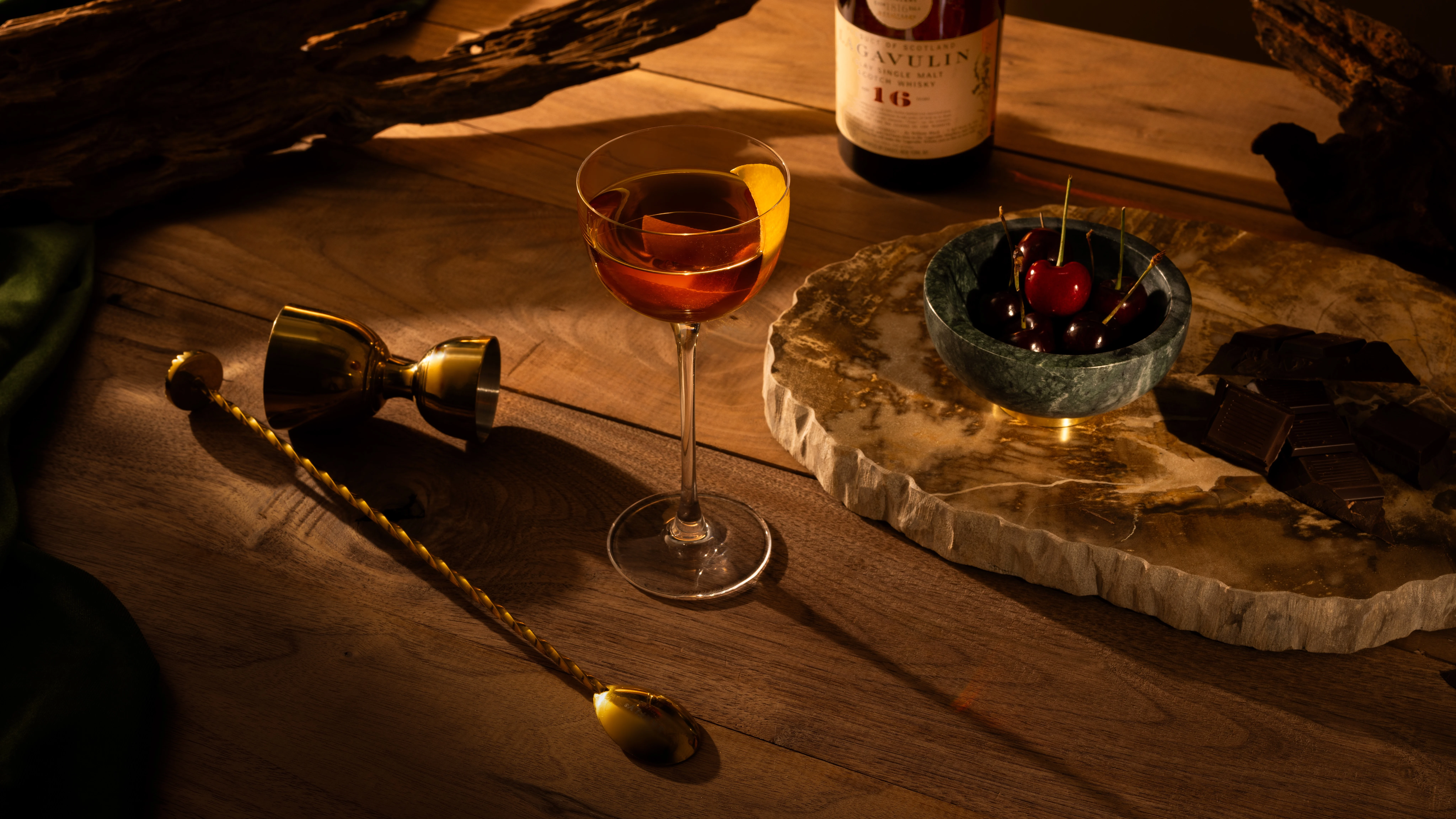 A Peaty Burns cocktail in a coupe glass on a wooden surface surrounded by cocktail-making equipment.