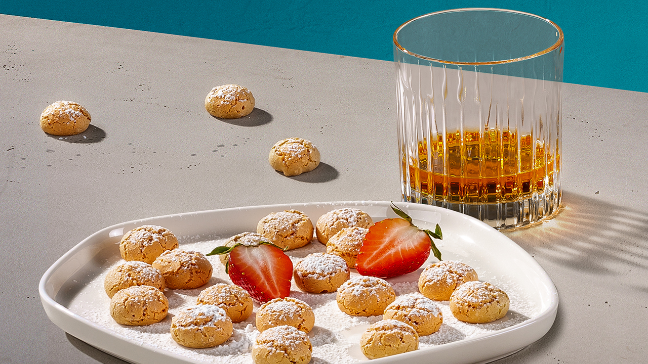 A glass of whisky next to a plate of powdered sugar-coated macaroons and sliced strawberries on a light gray surface.