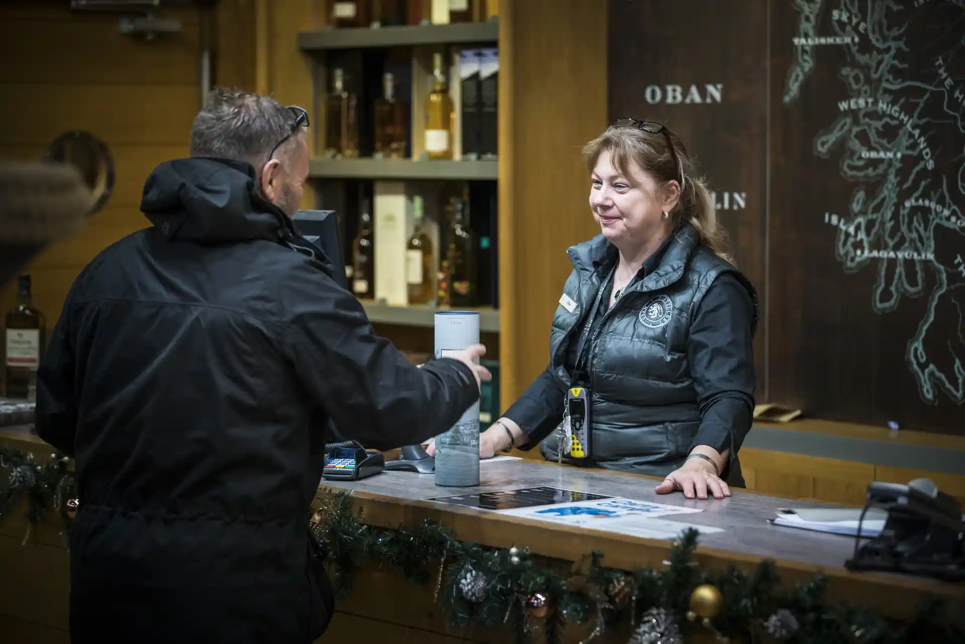 Een werknemer van de Oban distilleerderij staat achter een toonbank en bedient een klant die een fles Oban whisky koopt. Achter de medewerker staan planken met andere flessen whisky en een muur met een kaart van Schotland.