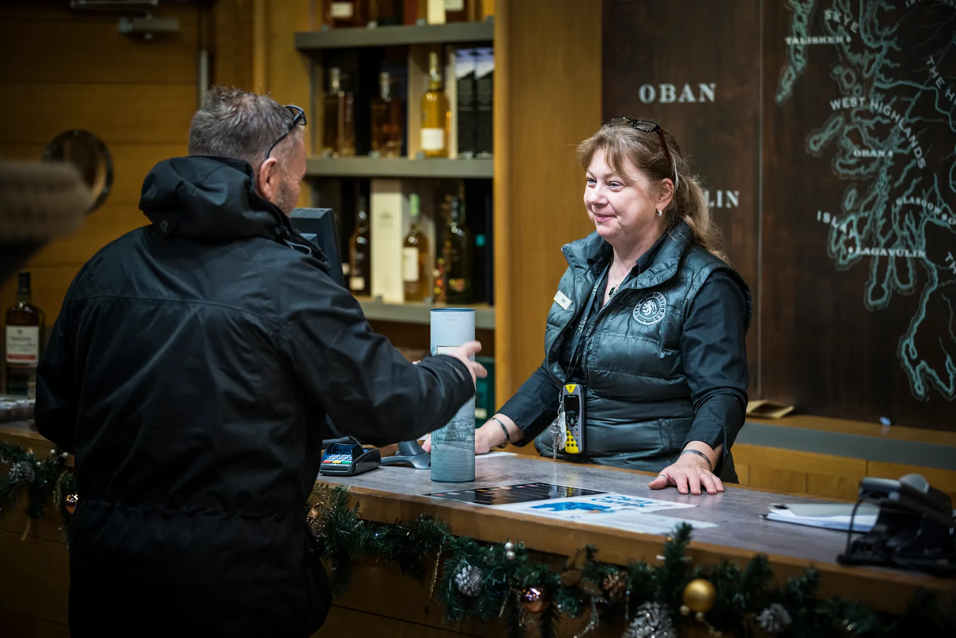 Ein Angestellter der Oban-Destillerie steht hinter einem Tresen und bedient einen Kunden, der eine Flasche Oban-Whisky kauft. Hinter dem Angestellten befinden sich Regale mit anderen Whiskyflaschen und eine Wand mit einer Karte von Schottland.
