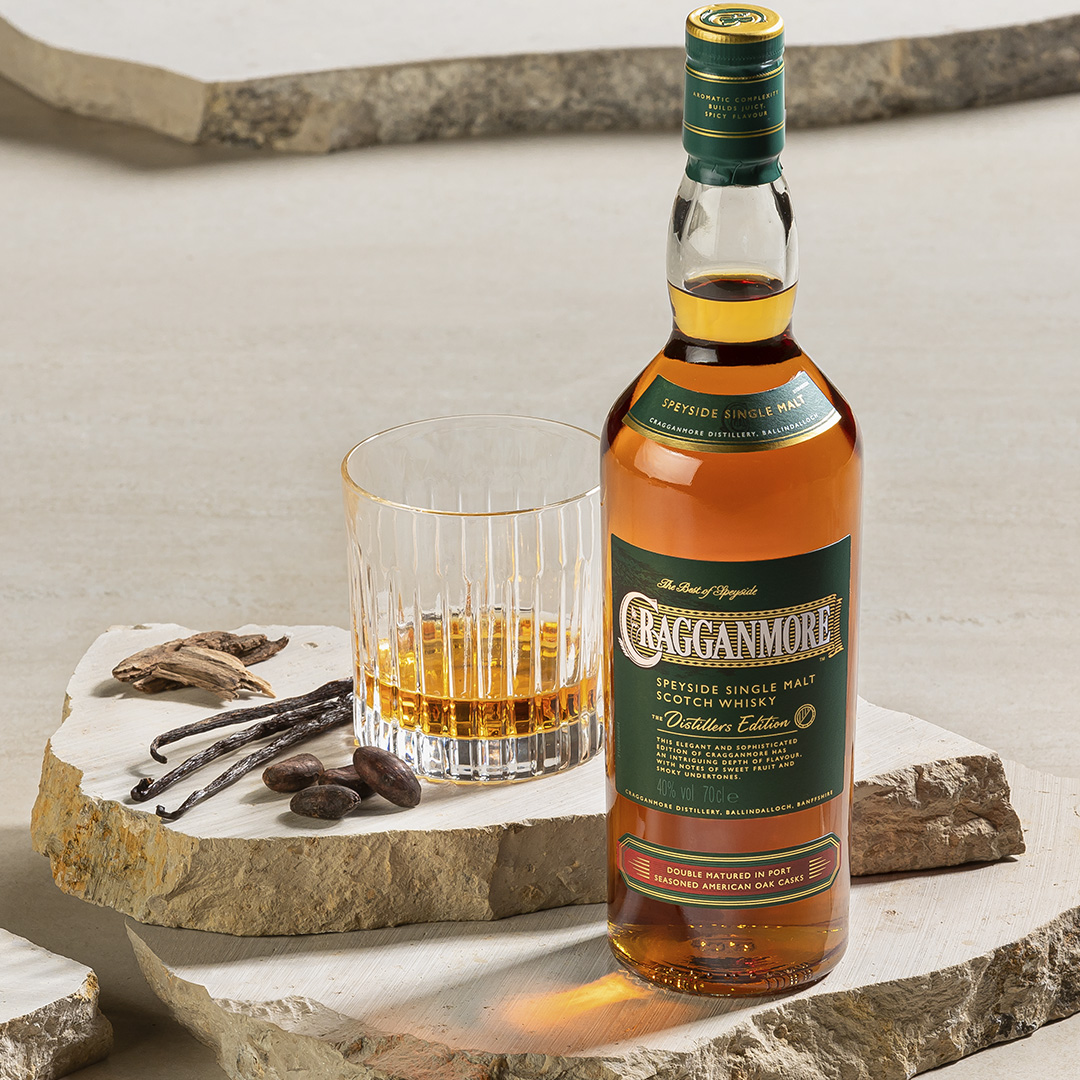 A bottle of Cragganmore Distillers Edition whisky sits on a marble slab. Next to it is a rocks glass with whisky, cacao beans and vanilla pods.