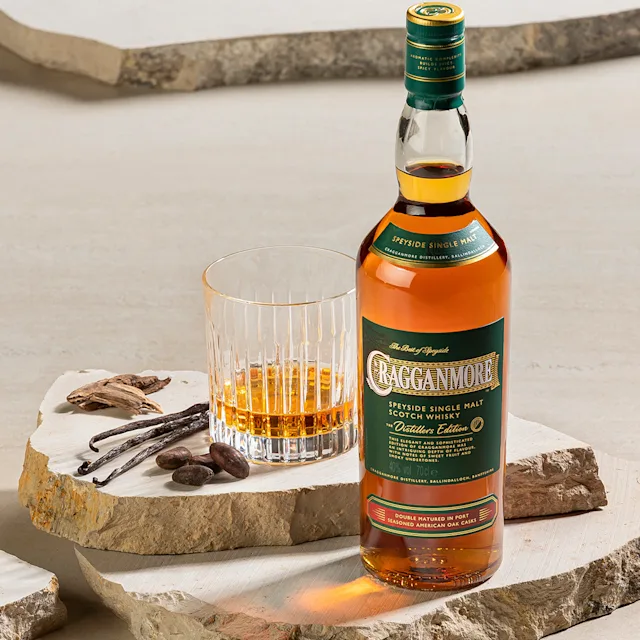 Een fles Cragganmore Distillers Edition whisky staat op een marmeren plaat. Ernaast staat een glas met whisky, cacaobonen en vanillestokjes.