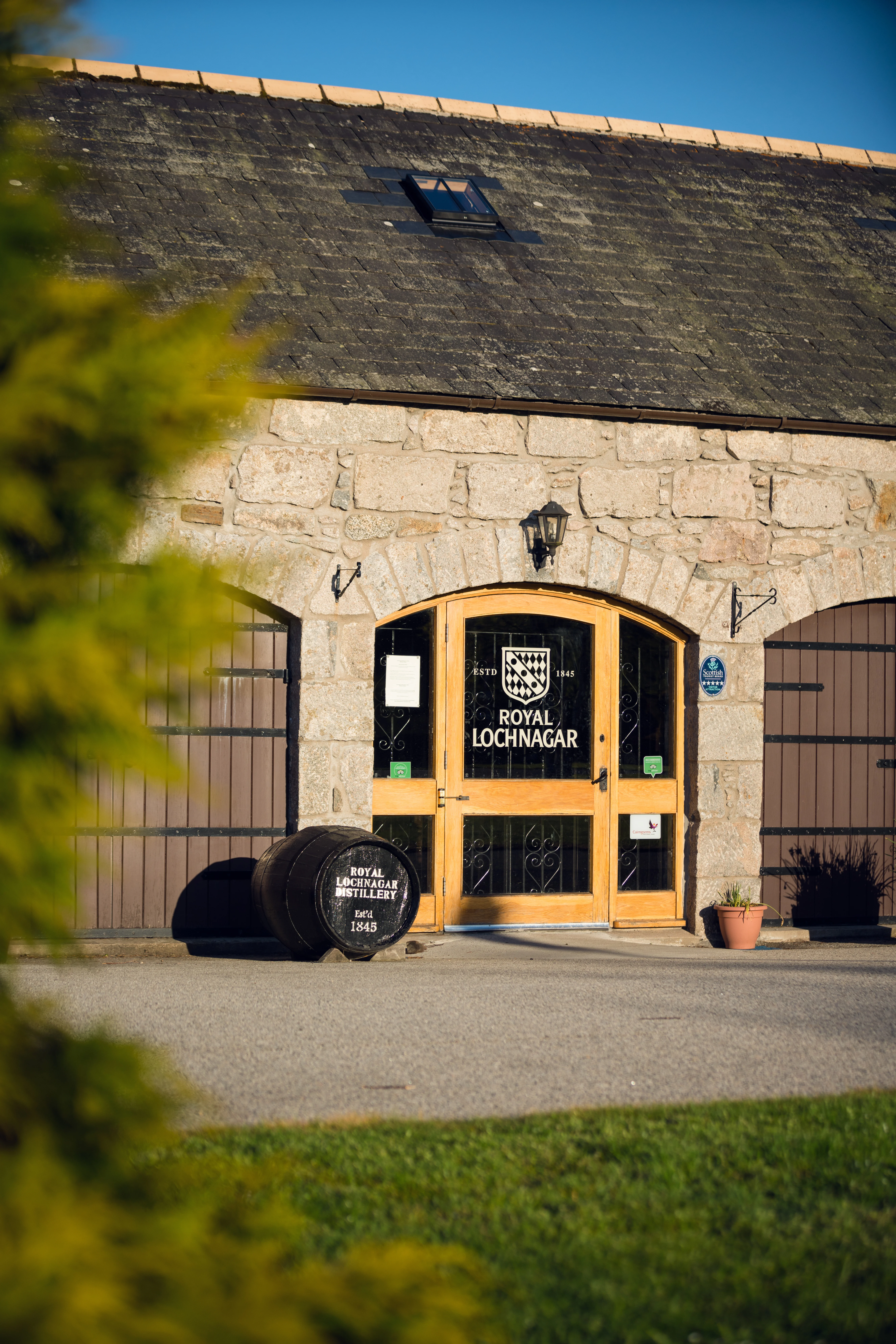 Wide-angle view of a Royal Lochnagar distillery with traditional stone buildings nestled among green hills and open highland countryside under a cloudy sky