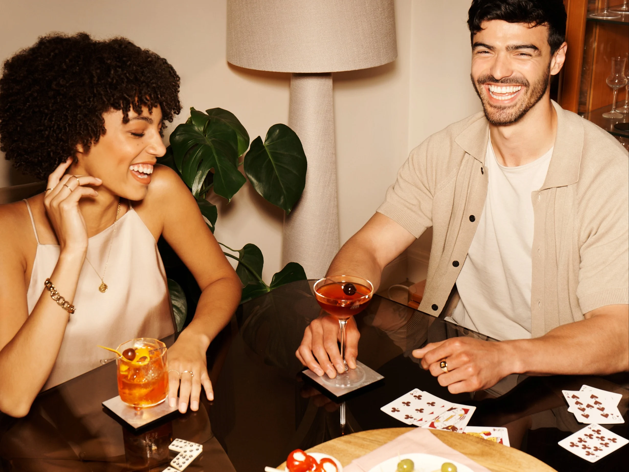 Zwei Freunde sitzen an einem Tisch und spielen Karten, in der Hand Cocktails.