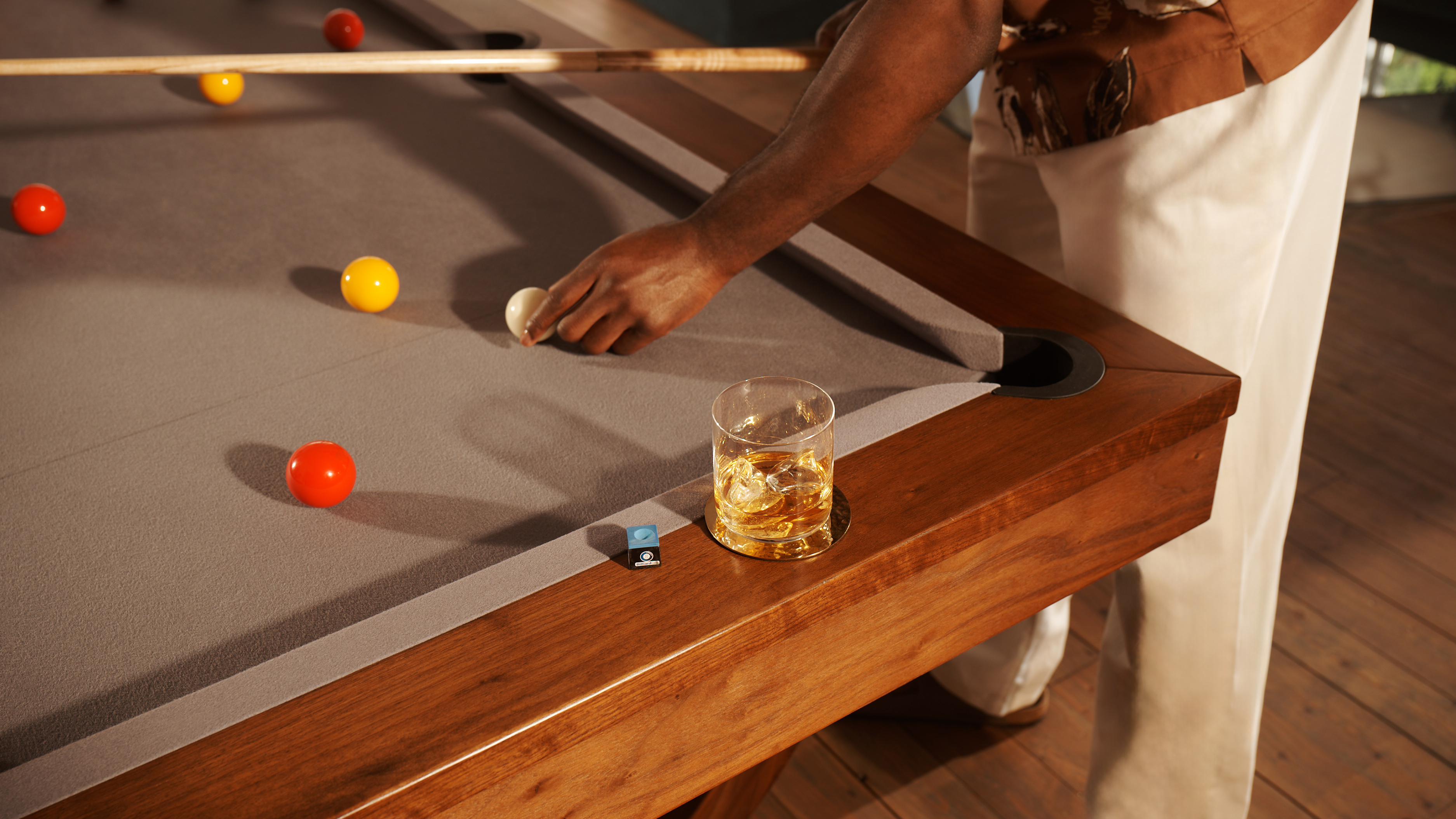 A man stands at a pool table, holding a pool cue and repositioning the white ball. A rocks glass of ice and whisky sits on the edge of the pool table.
