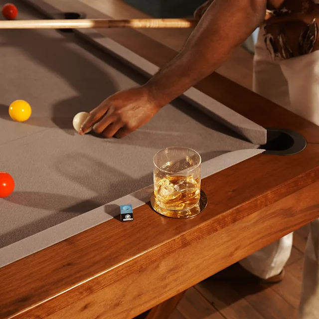 Ein Wasserglas mit Whisky und Eis steht auf dem Rand eines Billardtisches, auf dem mehrere Billardkugeln liegen. Jemand steht über dem Tisch, hält einen Queue und positioniert eine weiße Kugel neu.