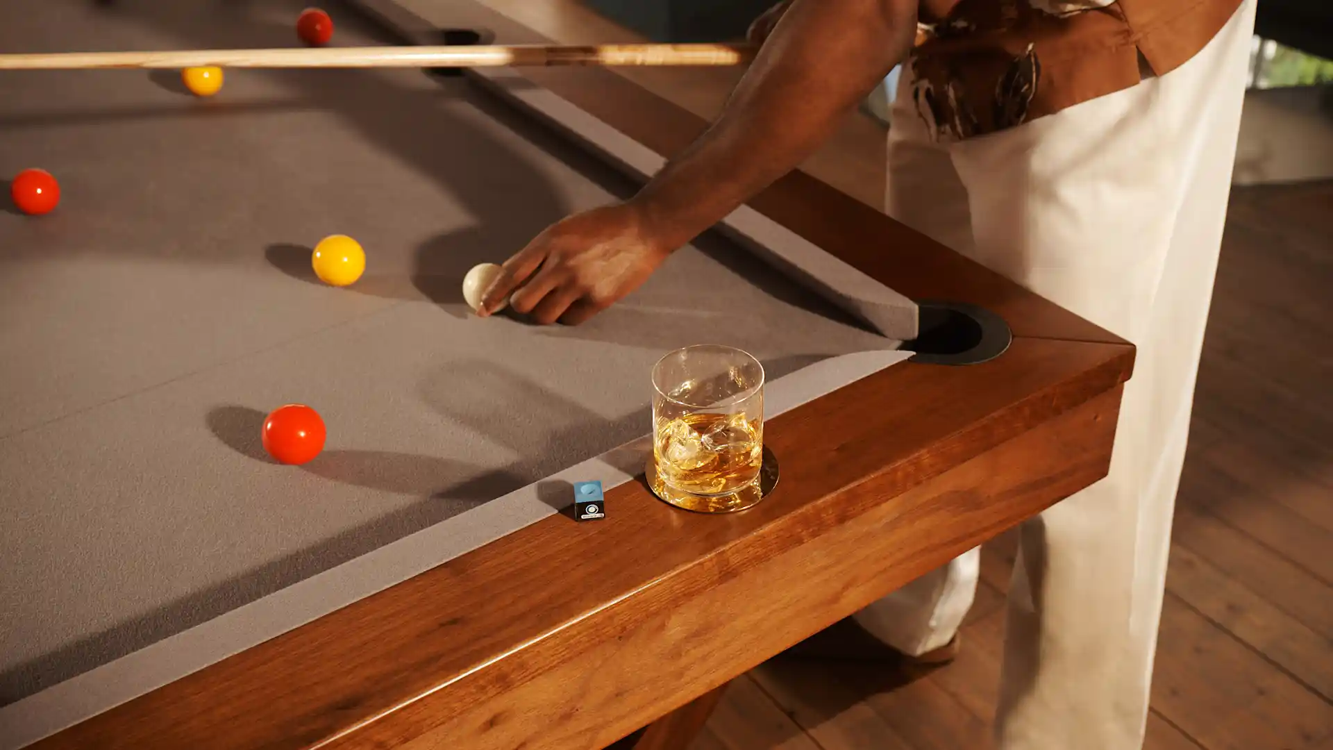 Een glas met whisky en ijs staat op de rand van een biljarttafel waarop verschillende biljartballen liggen. Iemand staat boven de tafel, houdt een keu vast en verplaatst een witte bal.