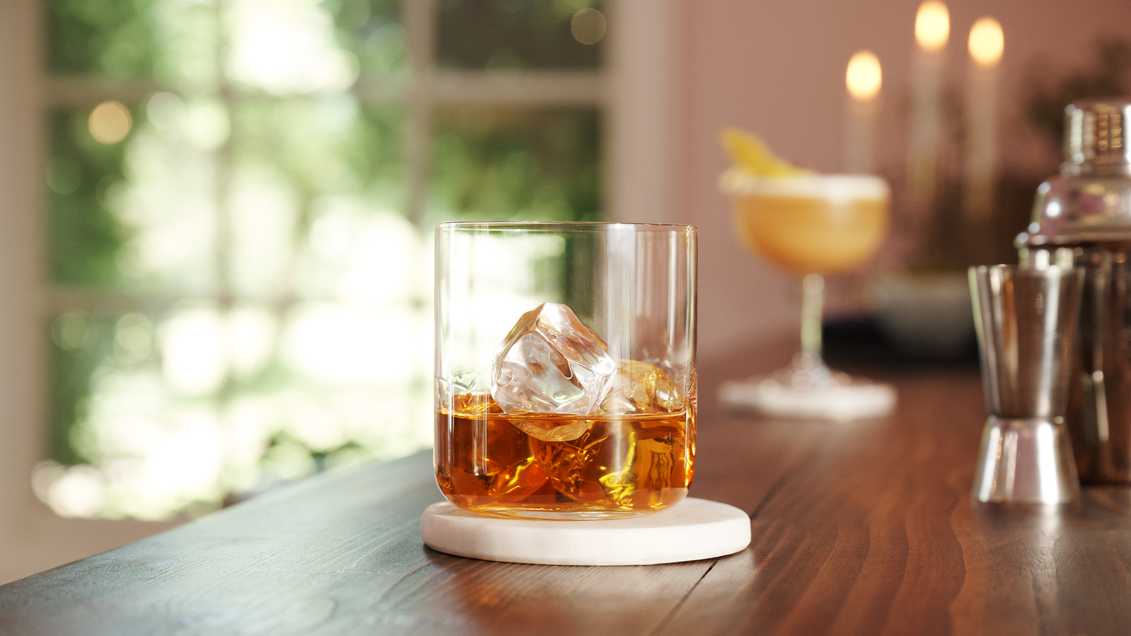 A rocks glass containing whisky and ice sits on a wooden surface with silver cocktail equipment and a cocktail in the background