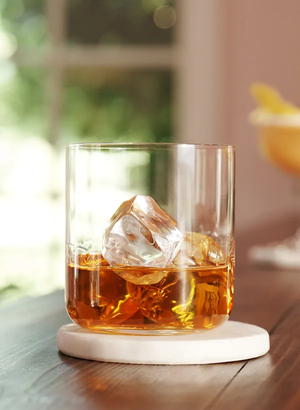 Een rocks glas met whisky en ijs staat op een houten oppervlak met zilveren cocktailspullen en een cocktail op de achtergrond