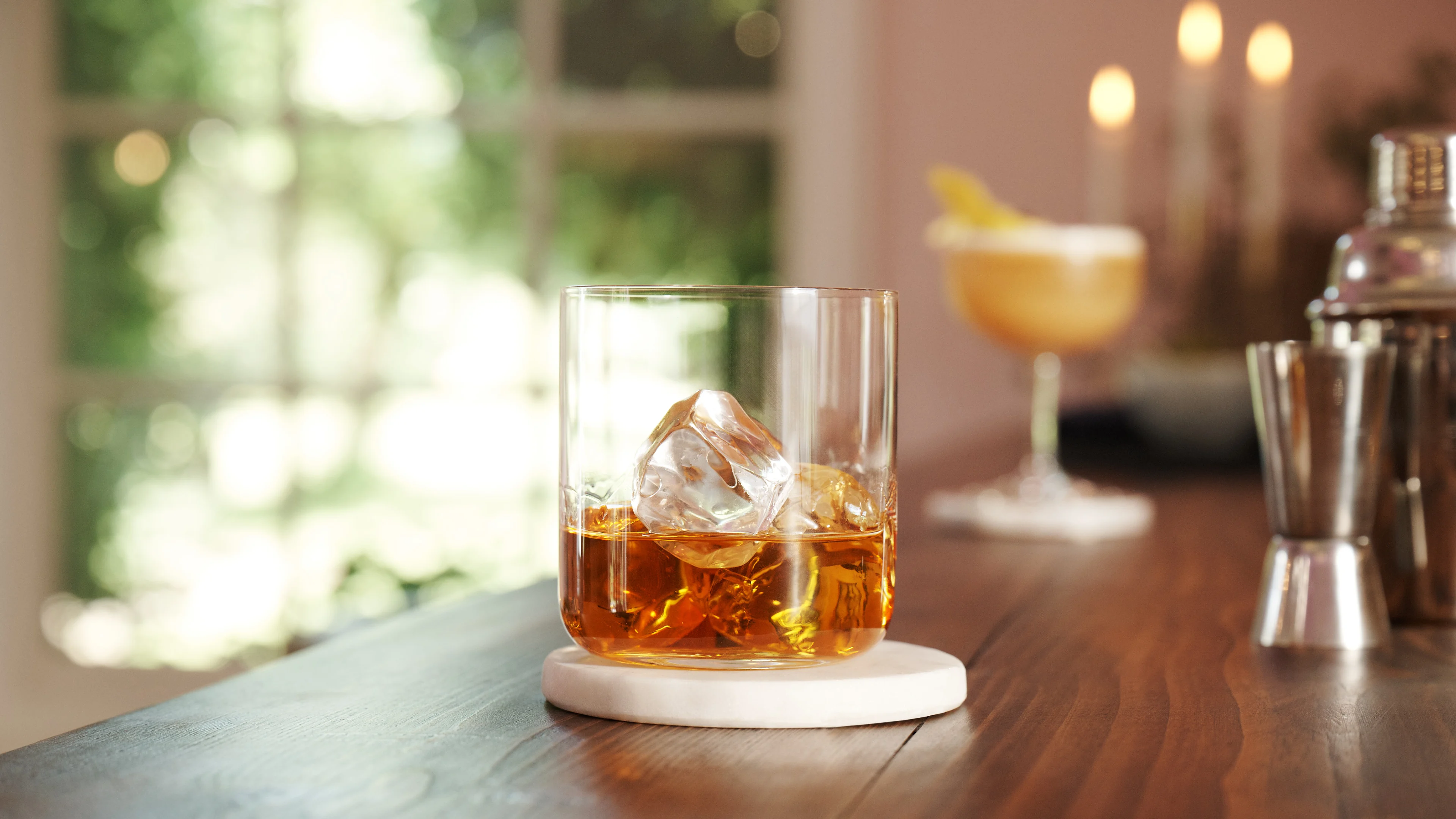A rocks glass containing whisky and ice sits on a wooden surface with silver cocktail equipment and a cocktail in the background