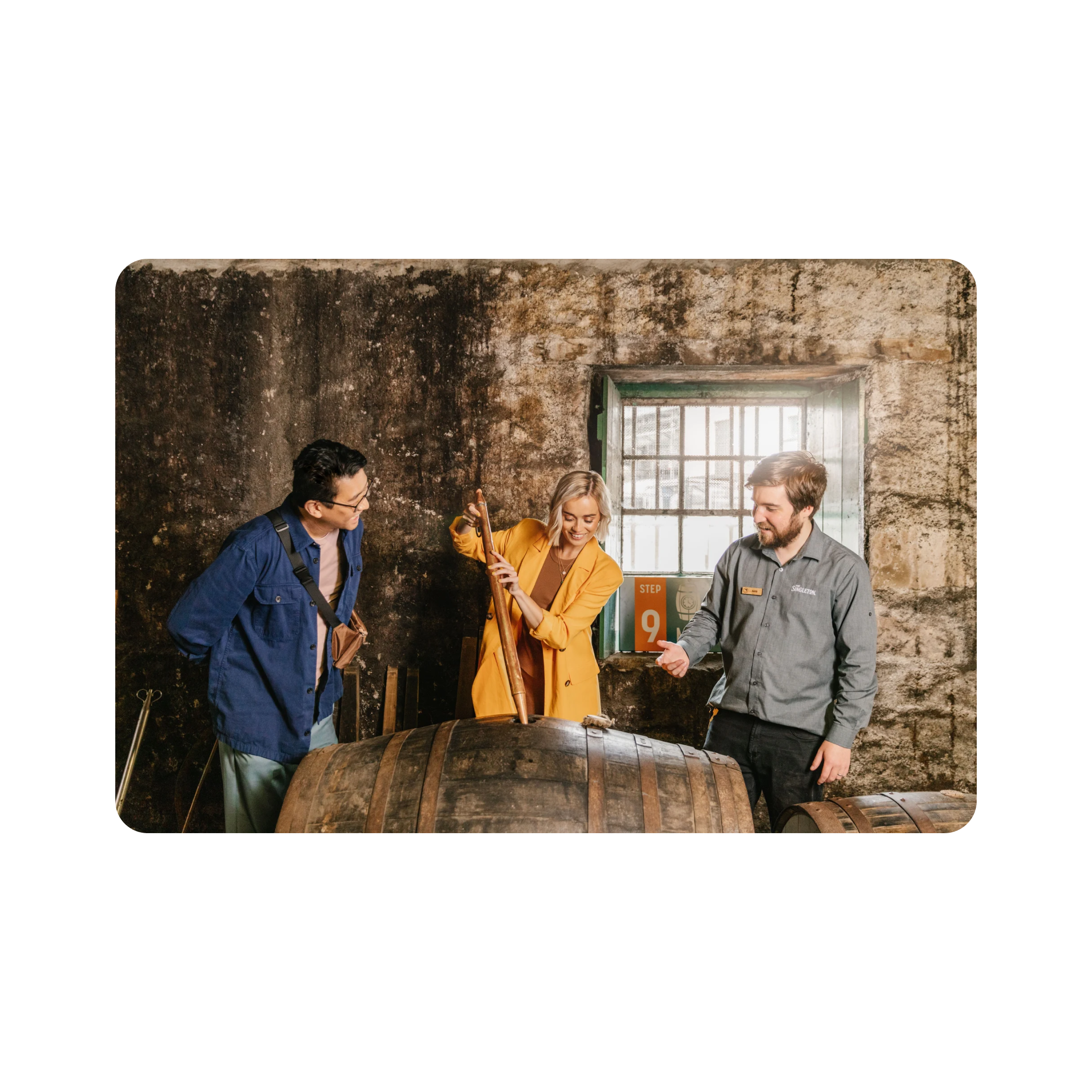 A lady taps into a new cask of The Singleton surrounded by an engaged observer and an informed guide . 