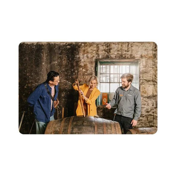 A lady taps into a new cask of The Singleton surrounded by an engaged observer and an informed guide .