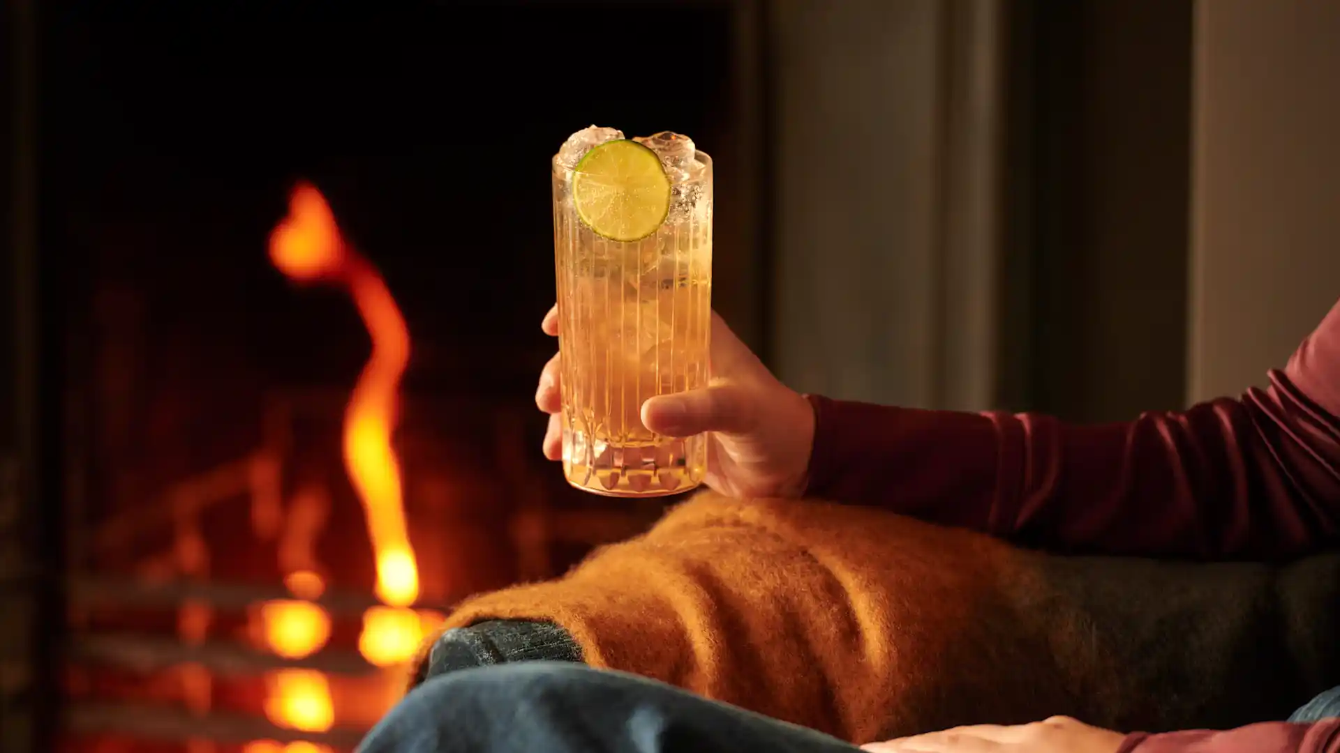 A woman sits in an armchair next to a roaring fire. She holds a whisky highball cocktail in her hands, which contains ice and a slice of lime.