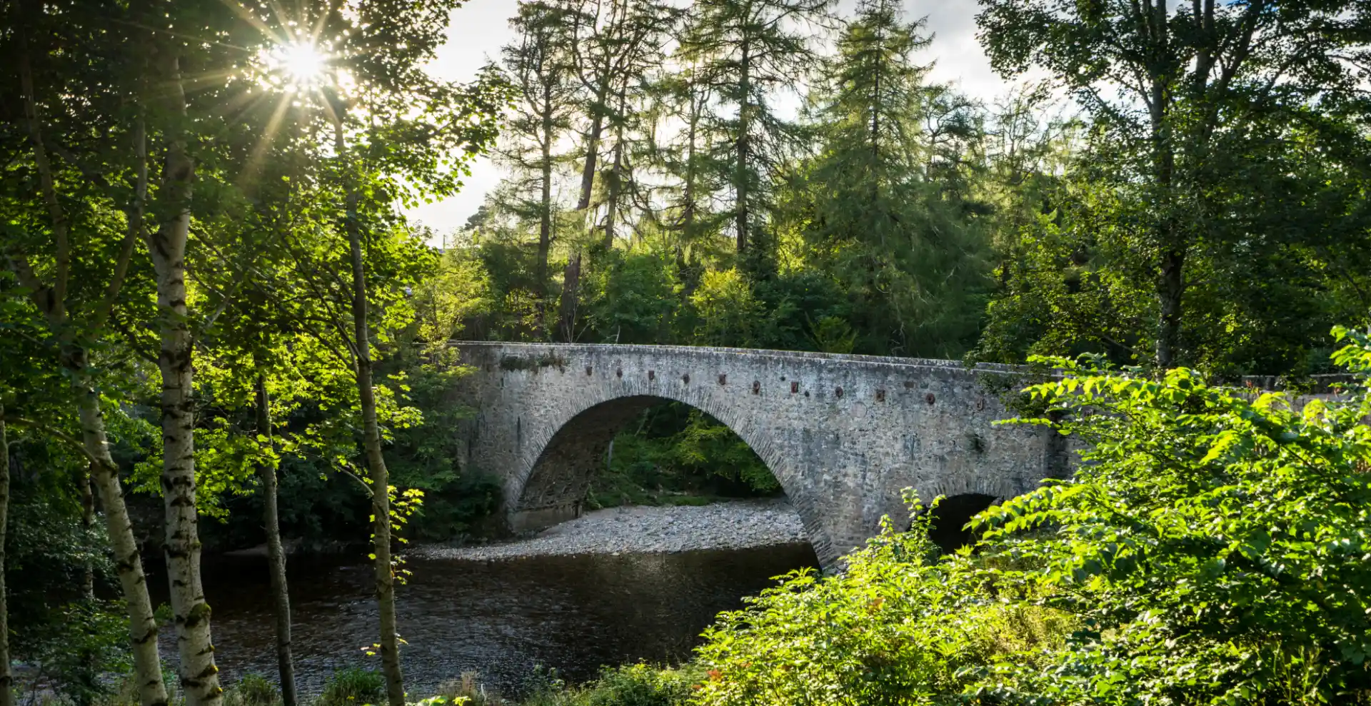 Eine graue Steinbrücke mit Bögen führt über einen kleinen Fluss. Um die Brücke herum gibt es viel grünes Laub und Bäume.