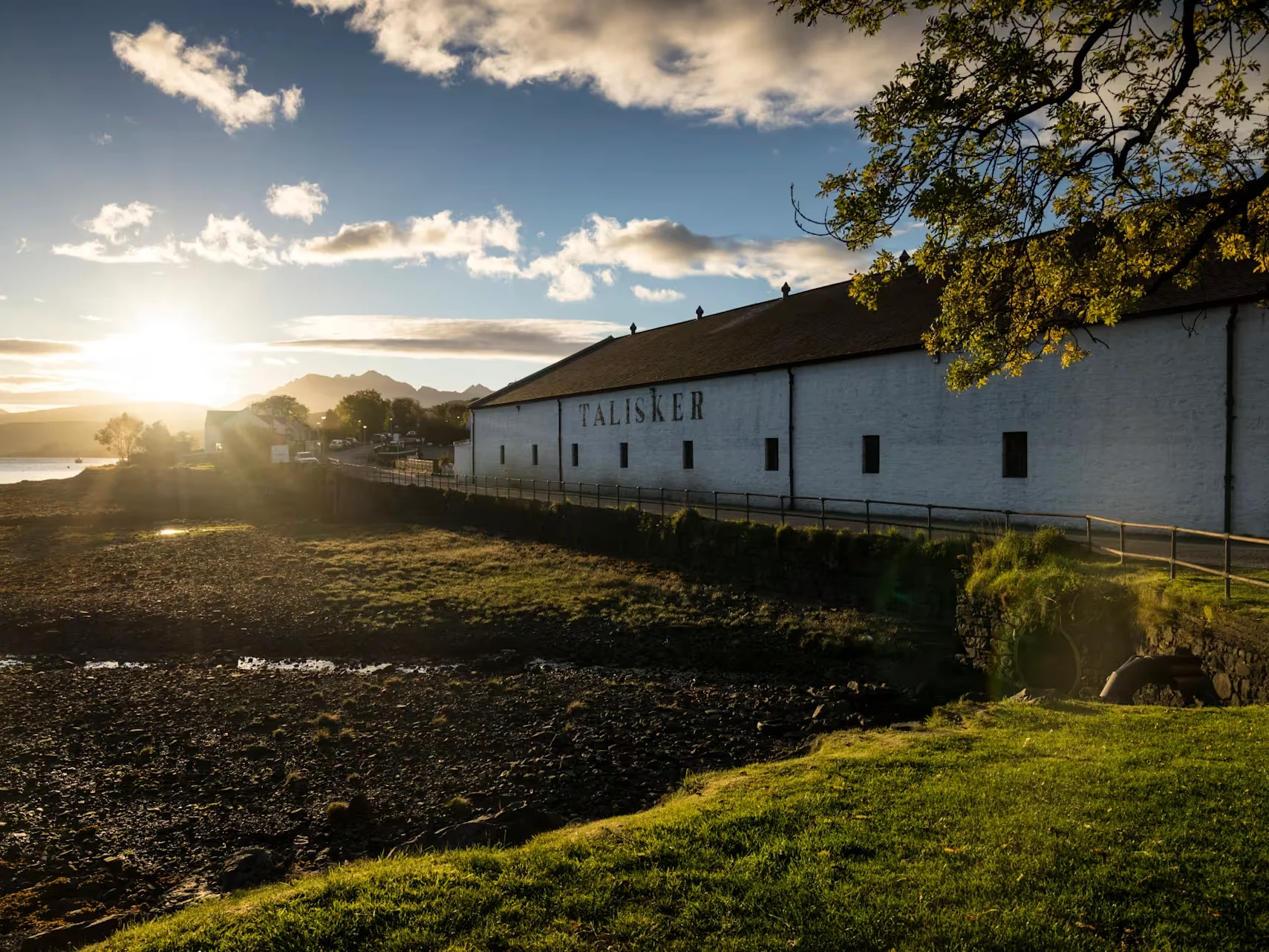Het witte gebouw van de Talisker distilleerderij staat op een groen stuk land, boven een kiezelstrand. De oever mondt uit in water en achter de horizon is een zonsondergang te zien.