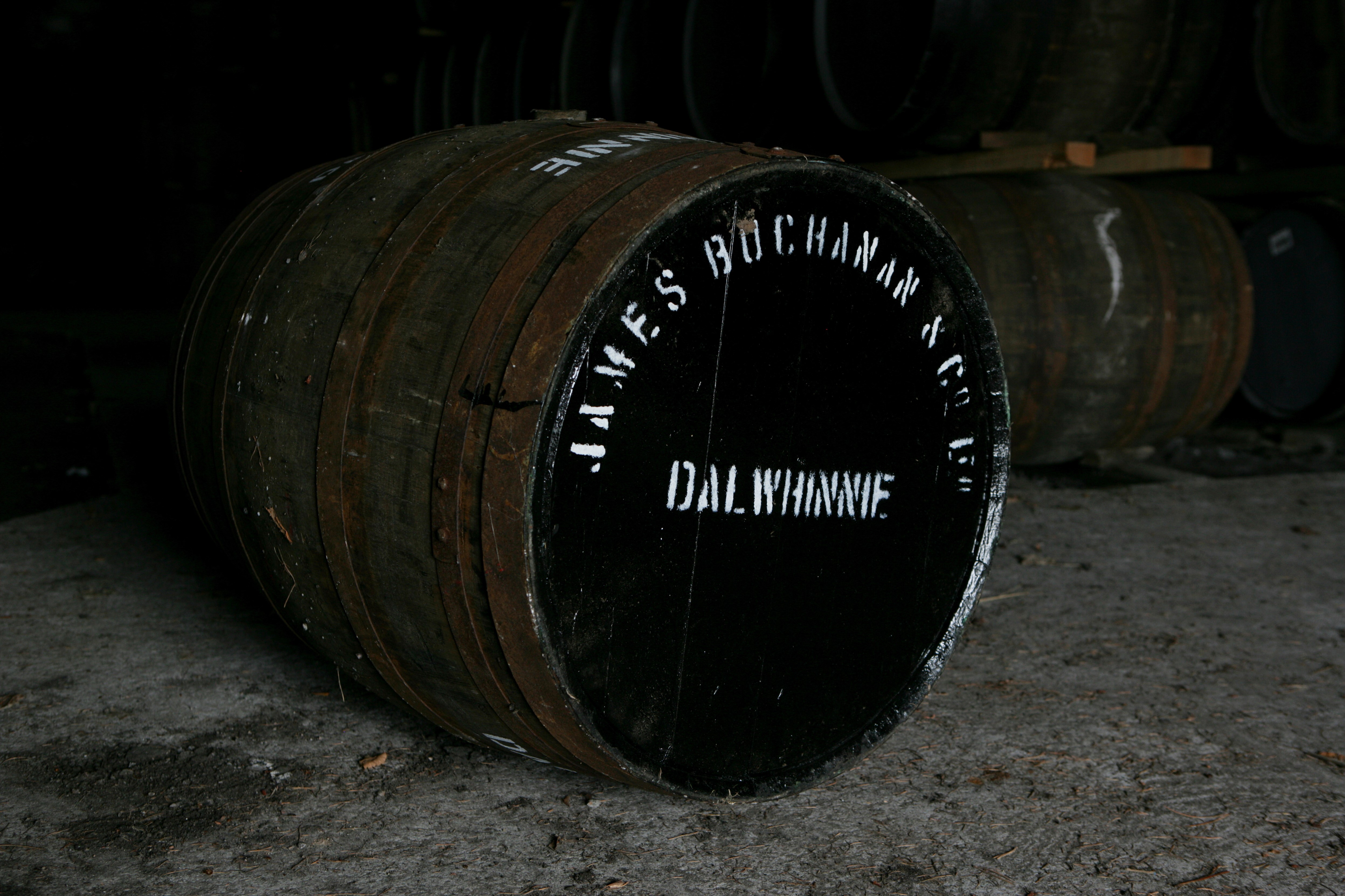 A wooden barrel lays on its side on a stone floor. The top of the barrel is painted black, and painted on with white letters are the words ‘James Buchanan & Co, Dalwhinnie’. There are other wooden barrels stacked in the background.
