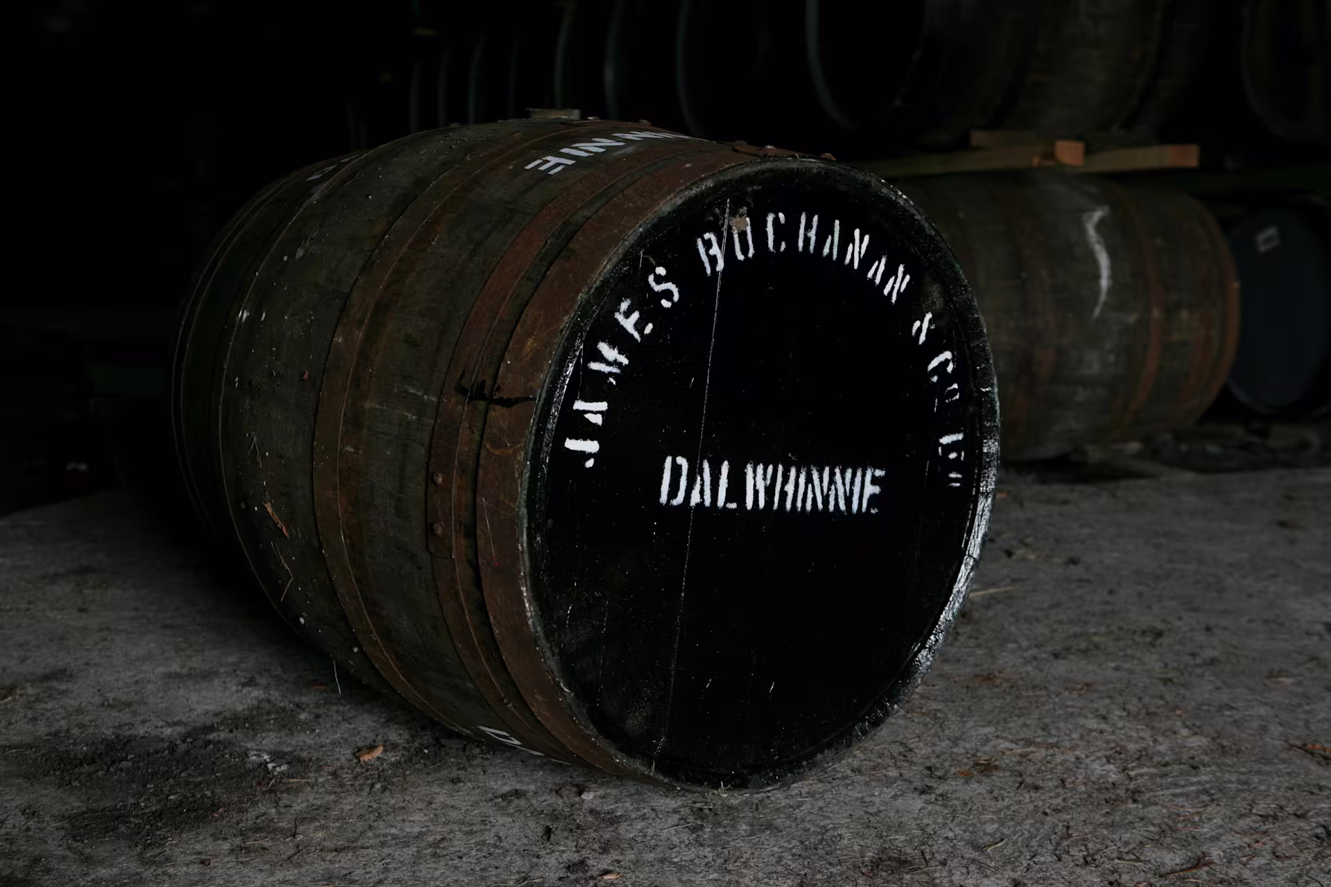 Ein Holzfass liegt auf der Seite auf einem Steinboden. Die Oberseite des Fasses ist schwarz gestrichen, und mit weißen Buchstaben sind die Worte "James Buchanan & Co, Dalwhinnie" aufgemalt. Im Hintergrund sind weitere Holzfässer gestapelt.