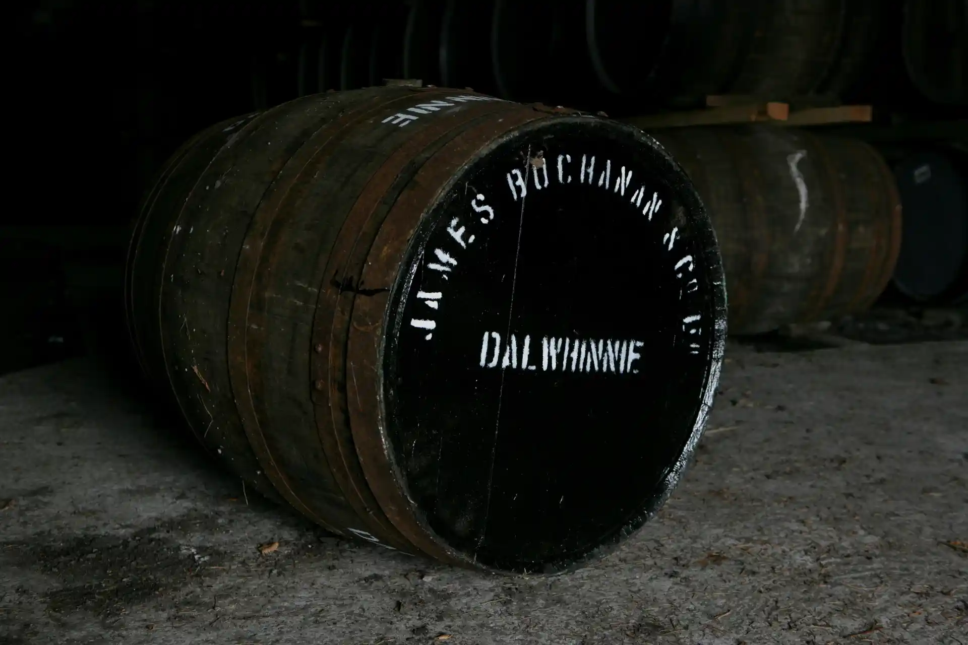 A wooden barrel lays on its side on a stone floor. The top of the barrel is painted black, and painted on with white letters are the words ‘James Buchanan & Co, Dalwhinnie’. There are other wooden barrels stacked in the background.