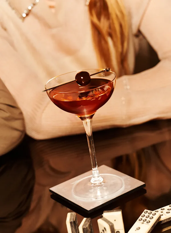 Three cocktails sit on a glass table. Two cocktails are Old Fashioneds with an orange peel and cherry garnish. The other cocktail is a Manhattan in a coupe glass with a skewered cherry garnish.