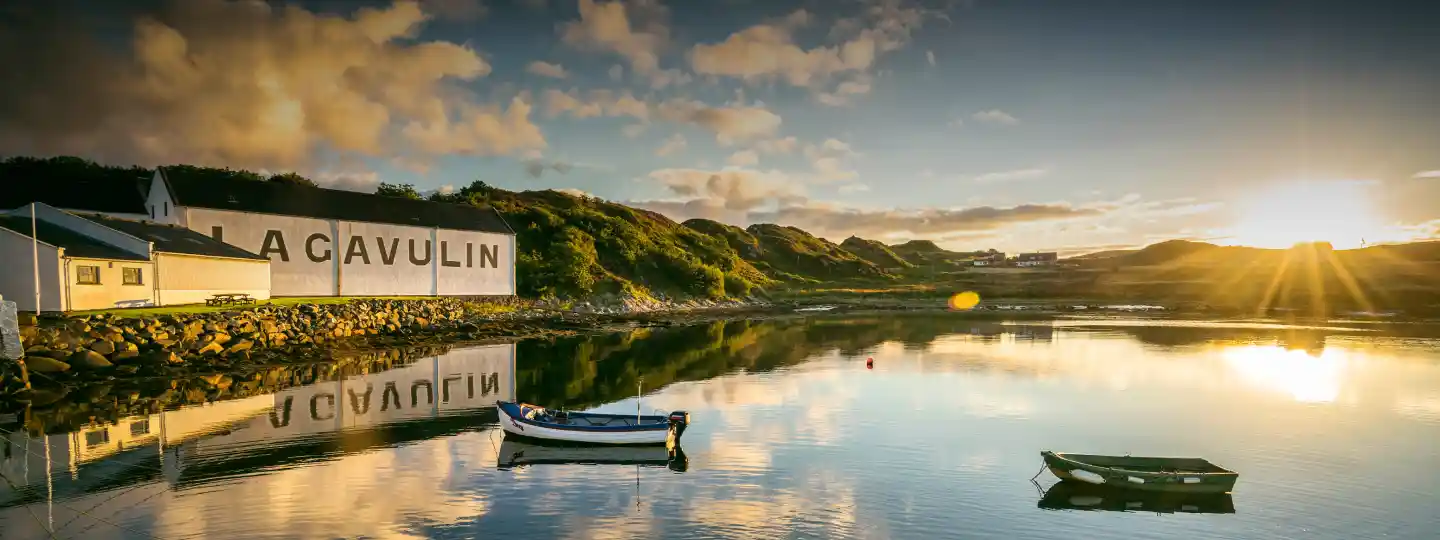 Two small boats sit on a body of water in front of a white building with the words Lagavulin painted onto the wall. Next to the building are grass-covered hills; the sky is blue and dotted with white clouds.