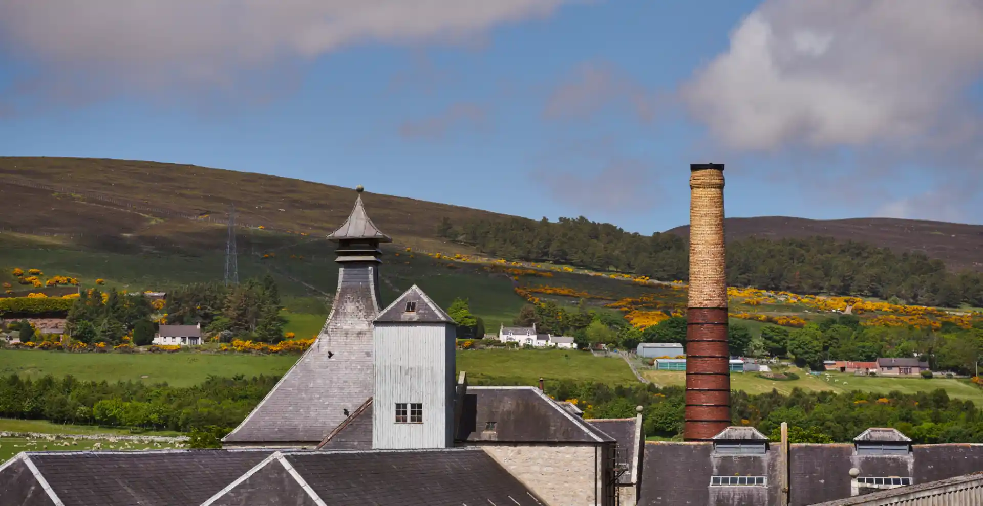 De Brora distilleerderij is van bovenaf te zien, met de gebouwen en het dak en de omliggende velden. Een persoon rolt een houten vat over een van de paden in de tuinen van de distilleerderij.  