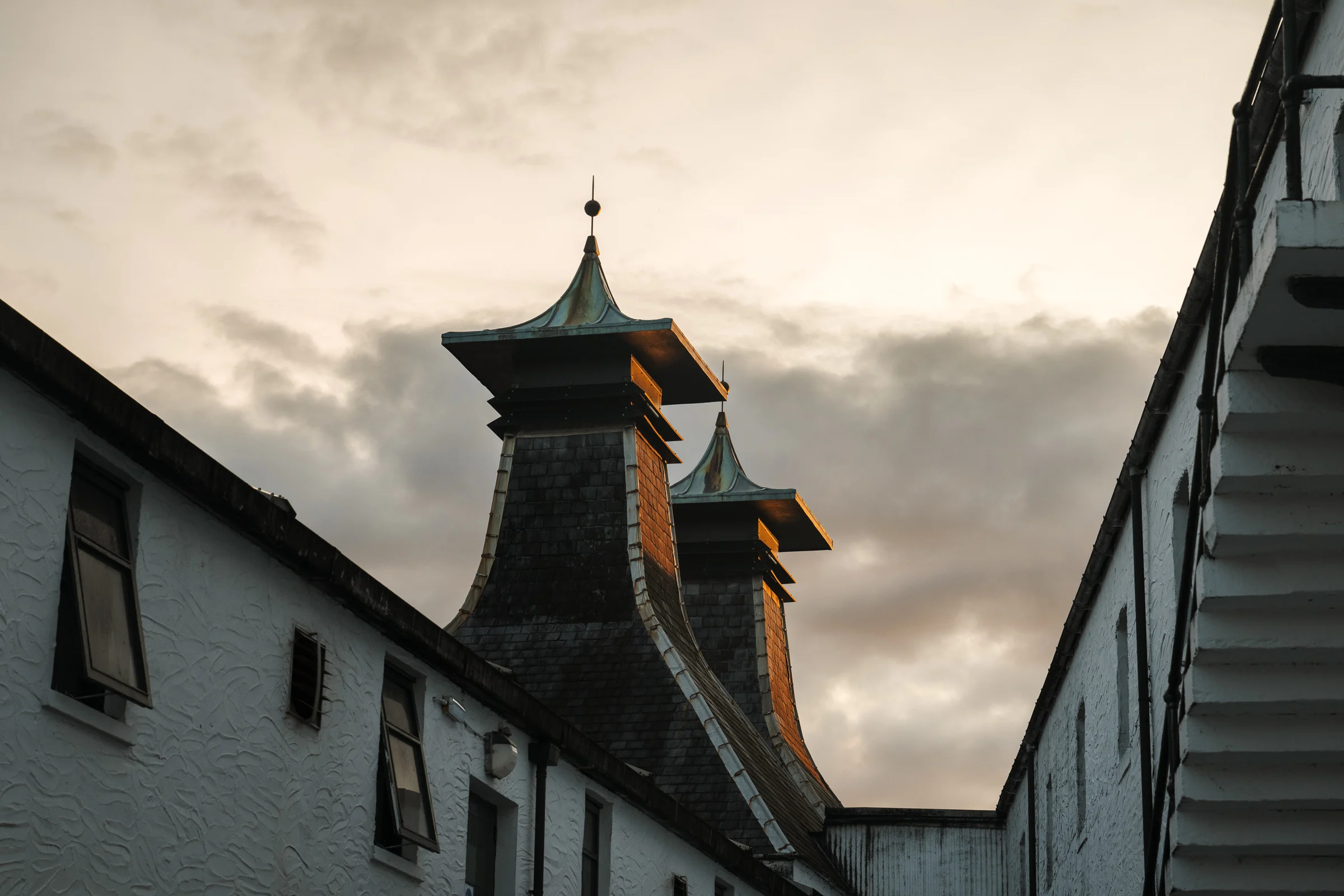 Traditional pagoda rooftops of an Islay whisky distillery rising above white-washed stone buildings against a dramatic cloudy sky at dusk.