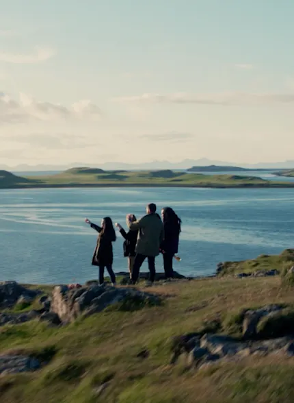 Vier mensen staan aan de rand van een klif en kijken uit over de zee ervoor en naar de horizon erachter. De klif is bedekt met rotsen en lange grassen die wapperen in de wind.