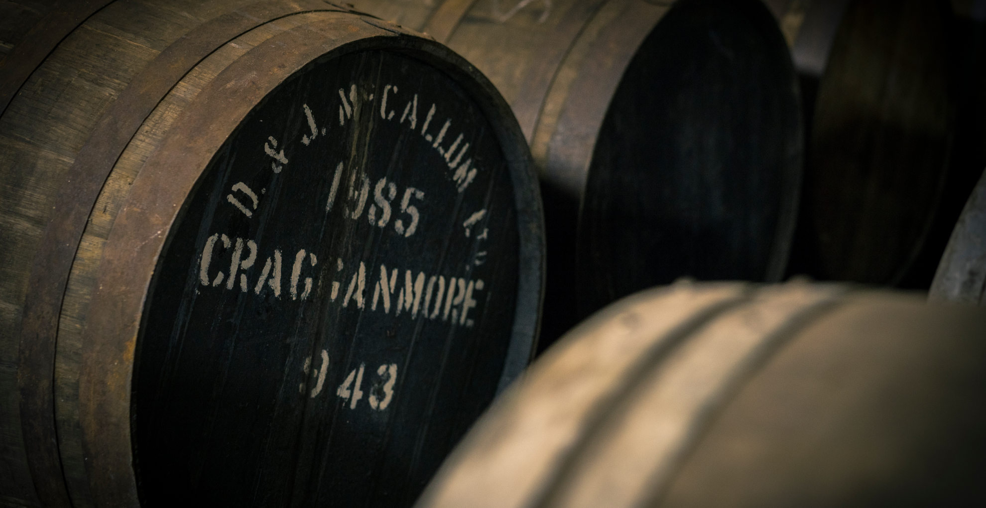 several wooden barrels sit in a row. One barrel has white writing on its front which says ‘D&J McCallum, 1985, Cragganmore, 943’.