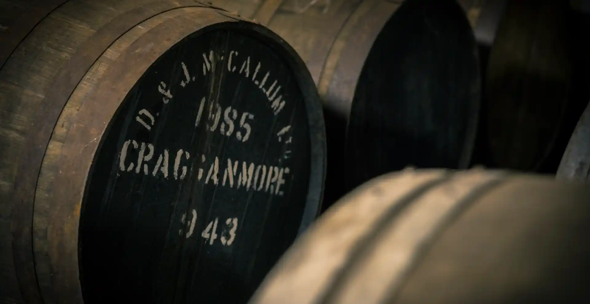 several wooden barrels sit in a row. One barrel has white writing on its front which says ‘D&J McCallum, 1985, Cragganmore, 943’.