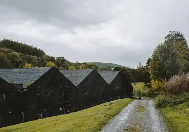 Several brick buildings sit in a row on a country path, surrounded by green fields, trees and a mountain.