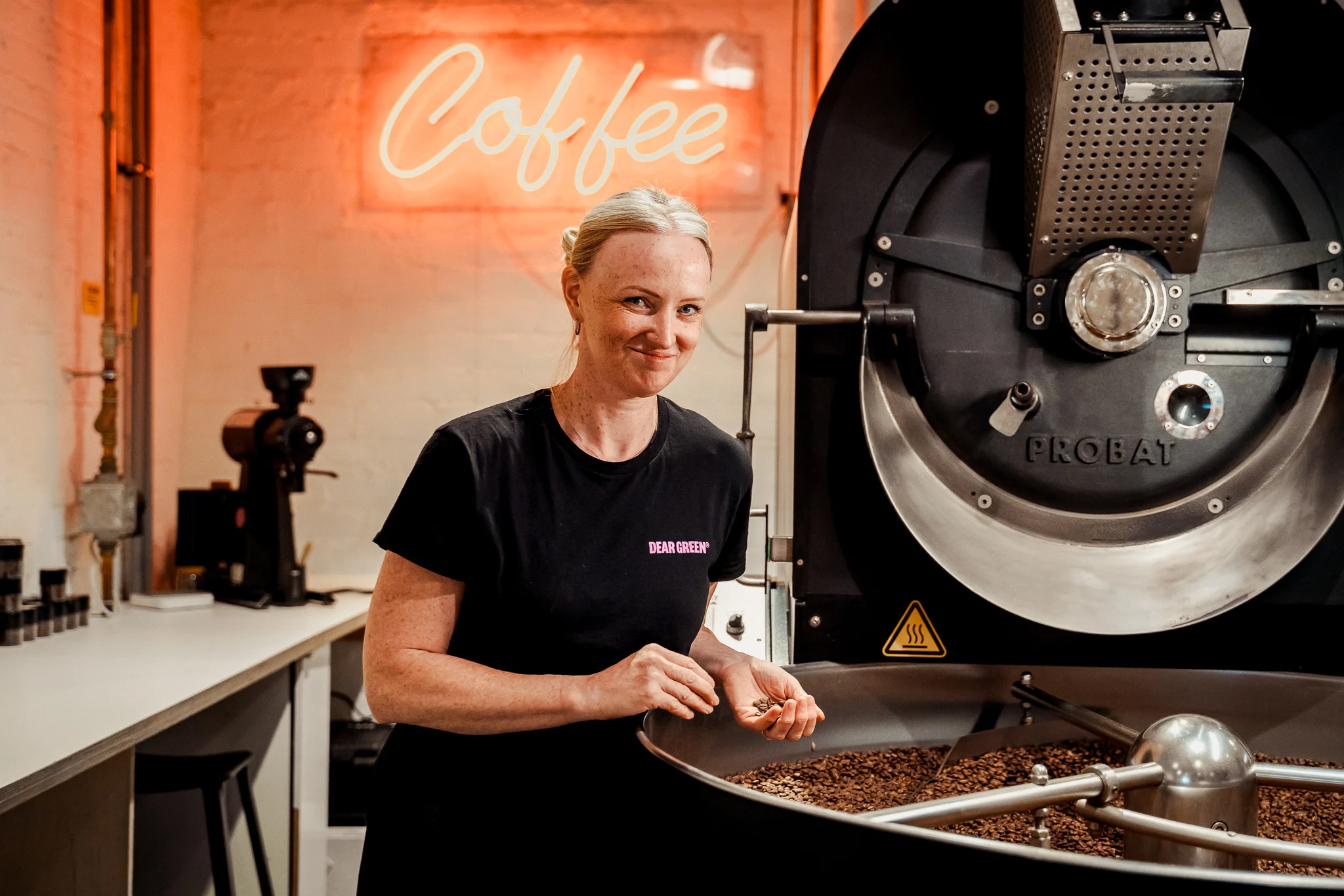 Lisa smiles whilst holding a handful of coffee brans standing in front of a neon sign saying "Coffee".