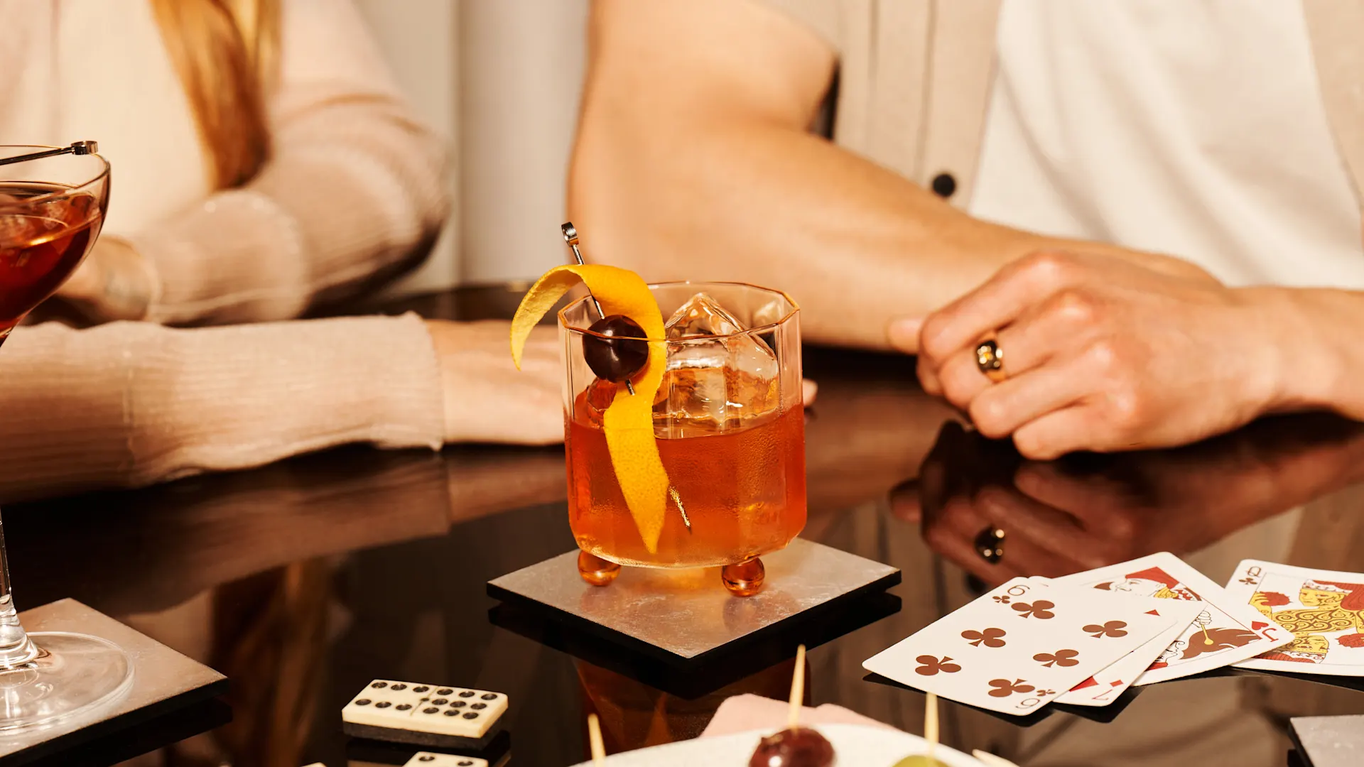 An Old Fashioned cocktail sits on a table surrounded by dominos and playing cards.