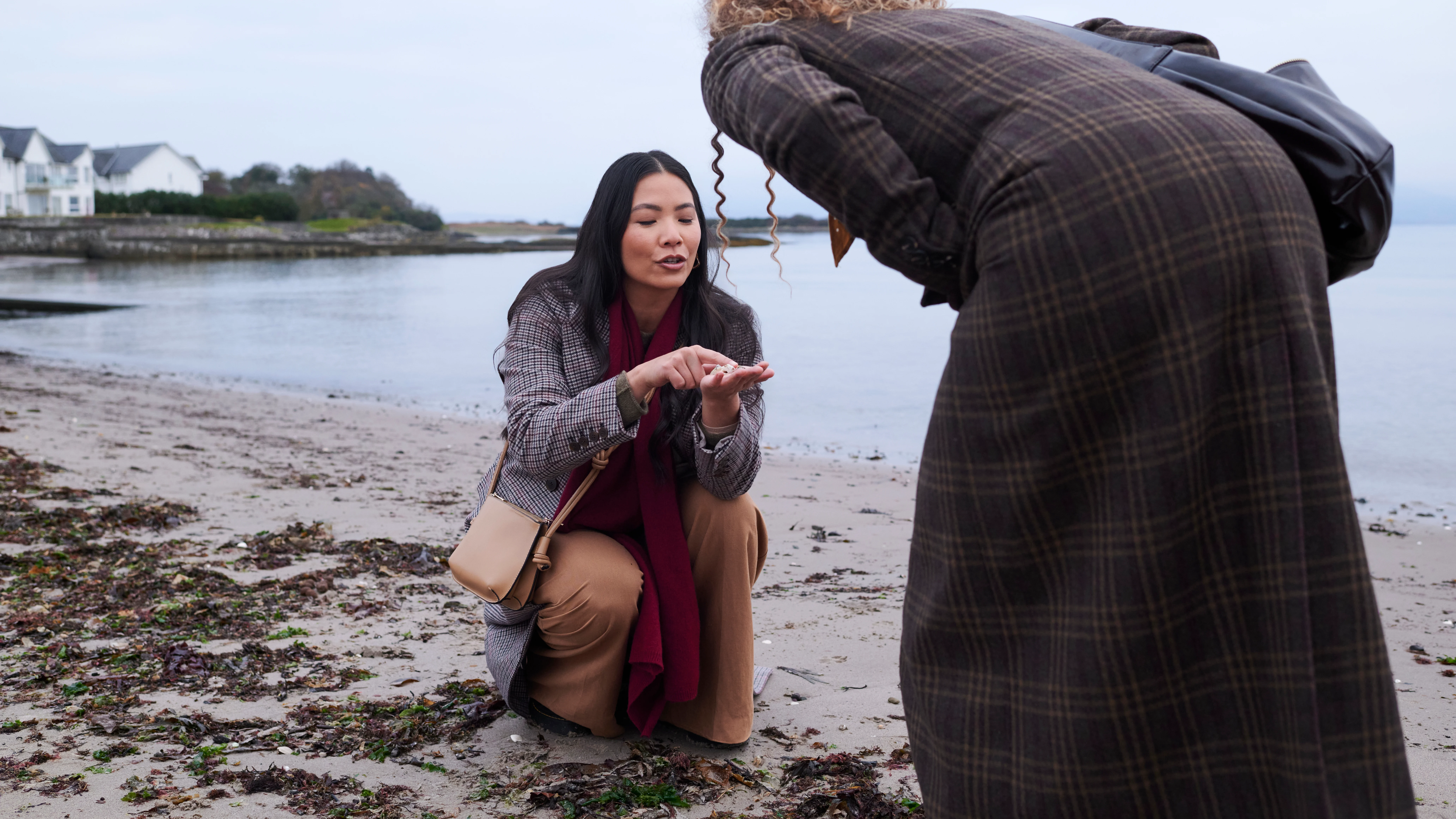 Two friends look at shells on a beach. One friend bends down to look at the shells another friend holds in her hands