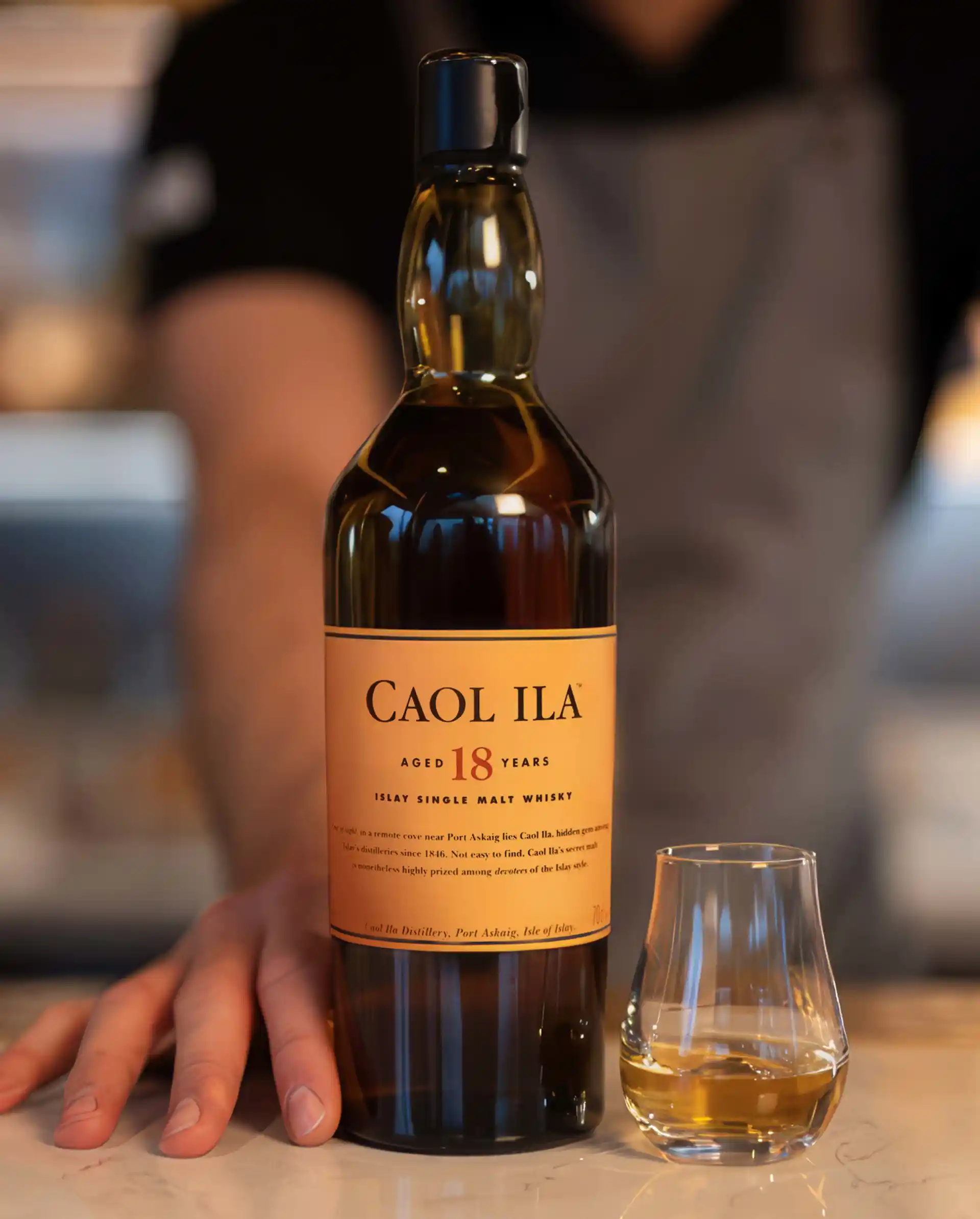 Een fles Caol Ila whisky staat op een marmeren tafelblad, naast een dramglas met whisky. Een persoon in een schort staat, in soft focus, op de achtergrond met zijn hand zachtjes rustend op de voet van de fles.