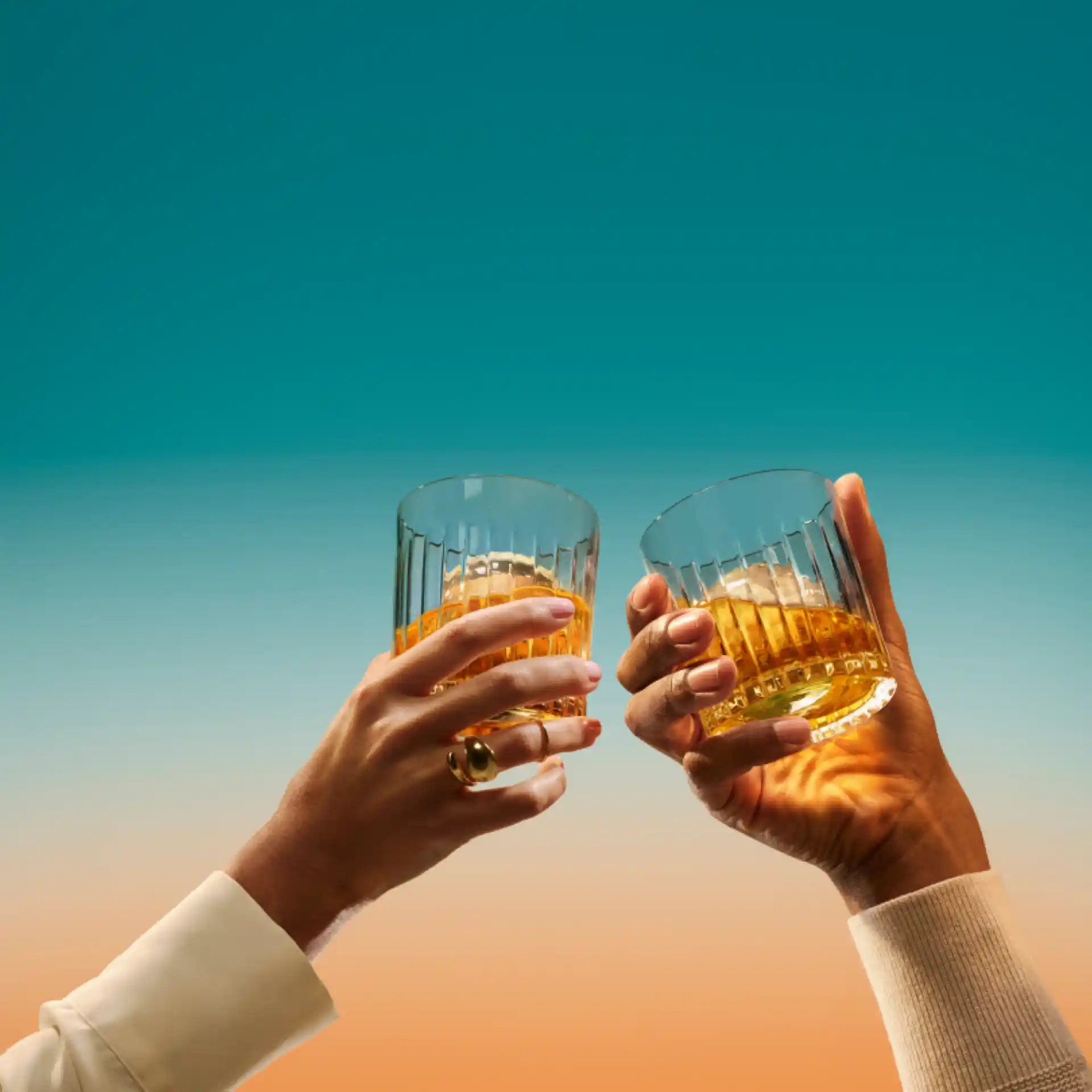 Twee handen houden elk een glas met ijs vast en brengen ze samen in een 'proost'-beweging. De glazen bevatten een groot ijsblokje en whisky.