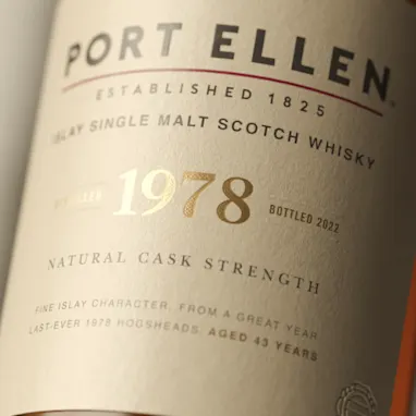 The white buildings of Port Ellen distillery overlook a body of water surrounded by a rocky shore. On one of the white walls, in large black writing, are the words ‘Port Ellen’.
