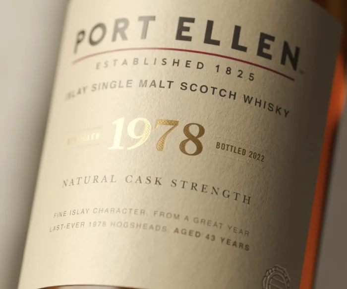 The white buildings of Port Ellen distillery overlook a body of water surrounded by a rocky shore. On one of the white walls, in large black writing, are the words ‘Port Ellen’.