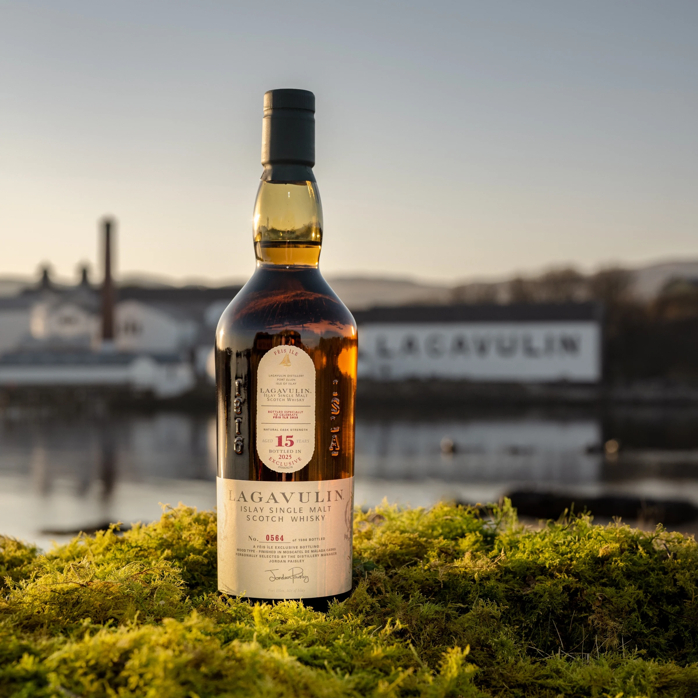 Lagavulin 15 Year Old Fèis Ìle 2025 Edition Flasche auf Moos mit der Lagavulin-Brennerei und der Küstenlinie im Hintergrund.