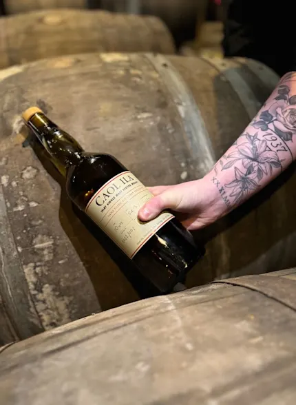 A bottle of Caol Ila is held between two wooden barrels. The person holds the bottle around the middle which shows the floral tattoos on their forearms.