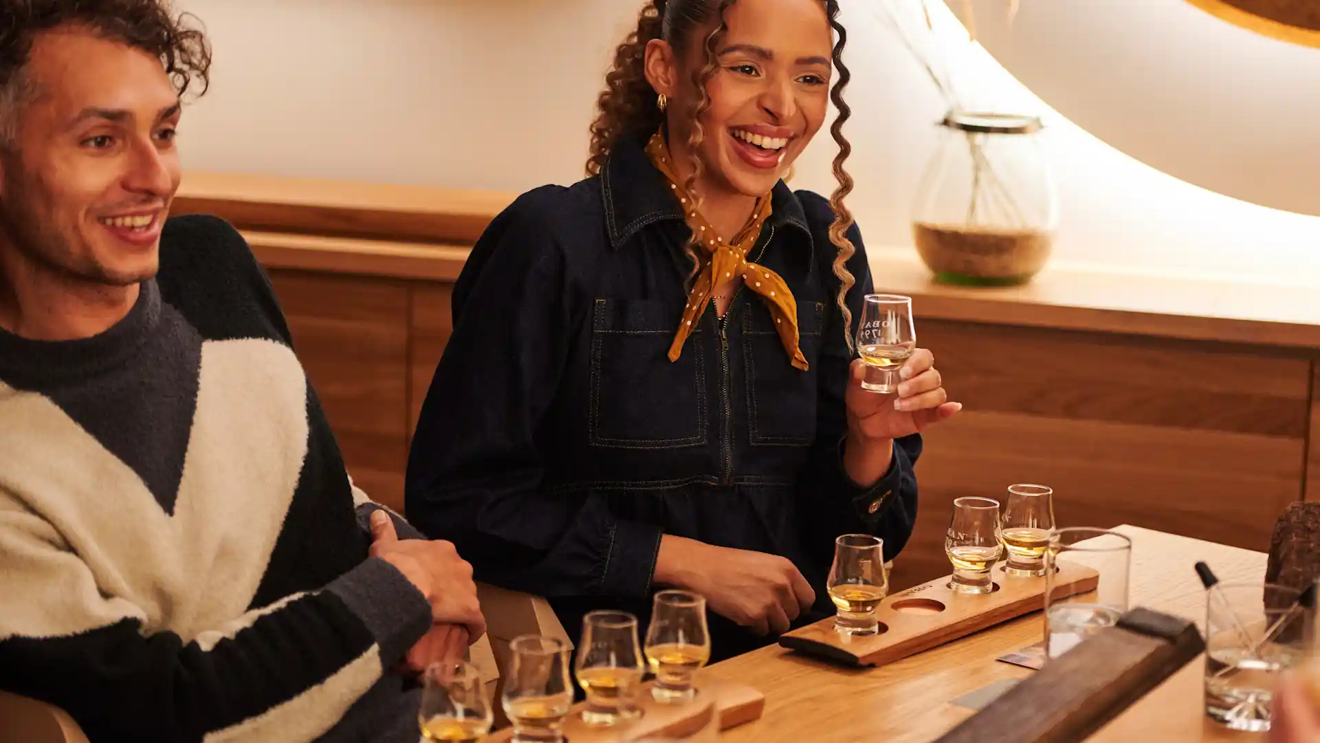 A man and a woman sit at a table enjoying a whisky tasting experience. In front of them are several small tasting glasses containing whisky.