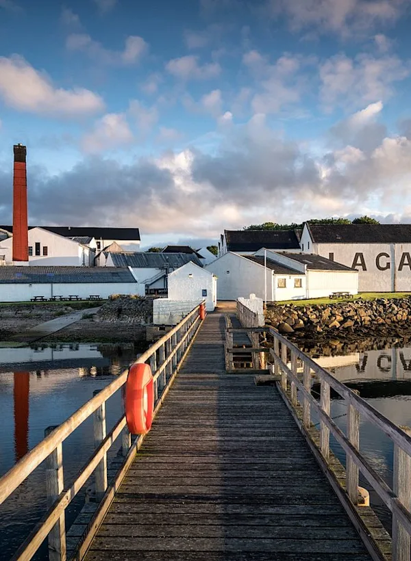 Eine Brennerei mit dem Schriftzug "Lagavulin" an der Seite, direkt am Wasser gelegen, mit einem Holzsteg, der zu ihr führt, unter einem teilweise bewölkten Himmel.