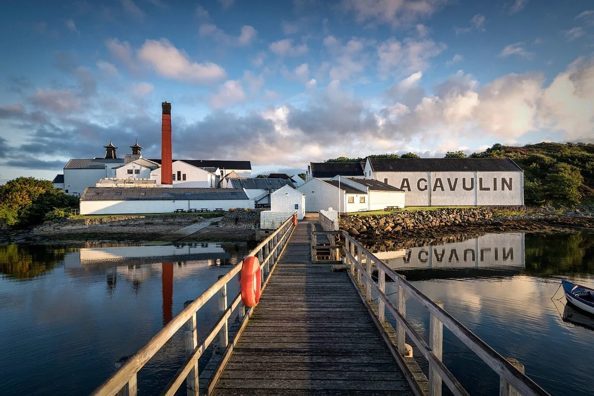 A distillery with "Lagavulin" written on the side, situated by the water with a wooden walkway leading to it, under a partly cloudy sky.