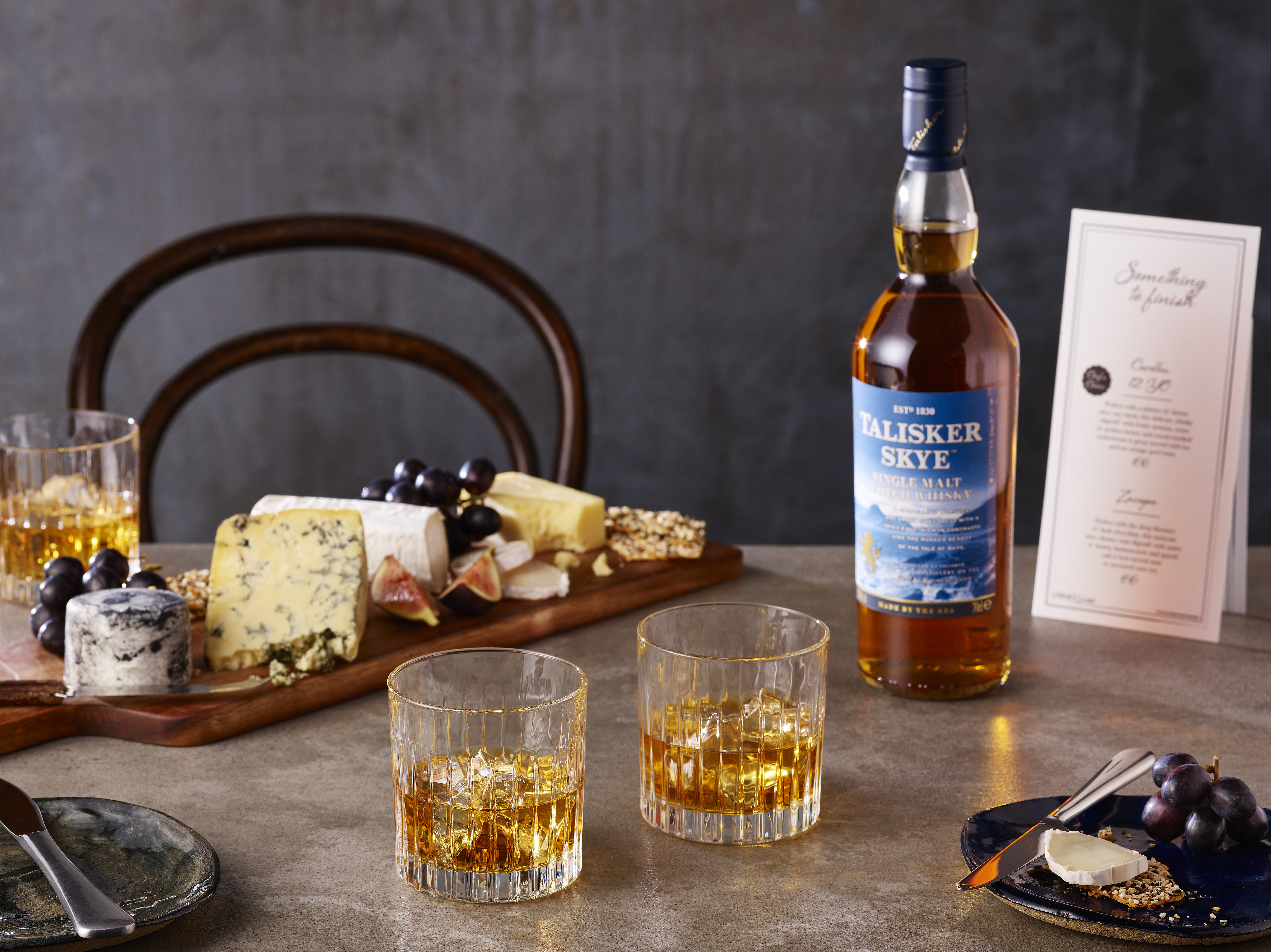 Een fles Talisker Skye staat op een tafel, naast twee glazen met ijs en whisky en een houten kaasplank met verschillende kazen en vruchten.