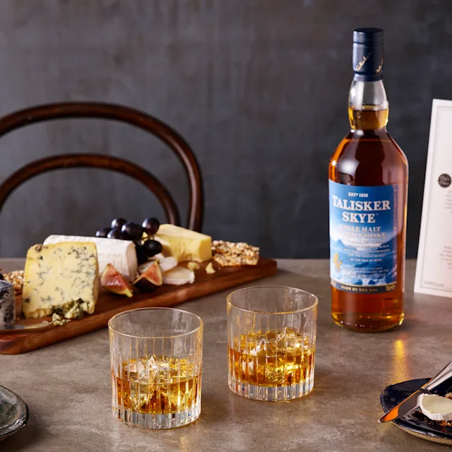 A bottle of Talisker Skye sits on a table, next to two rocks glasses containing whisky and ice, and a wooden cheeseboard displaying assorted cheeses and fruits.
