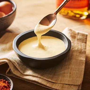 A small black dish containing a creamy coloured sauce sits on a table, surrounded by a bottle of bourbon, eggs and some spices. A spoon dips into the sauce.