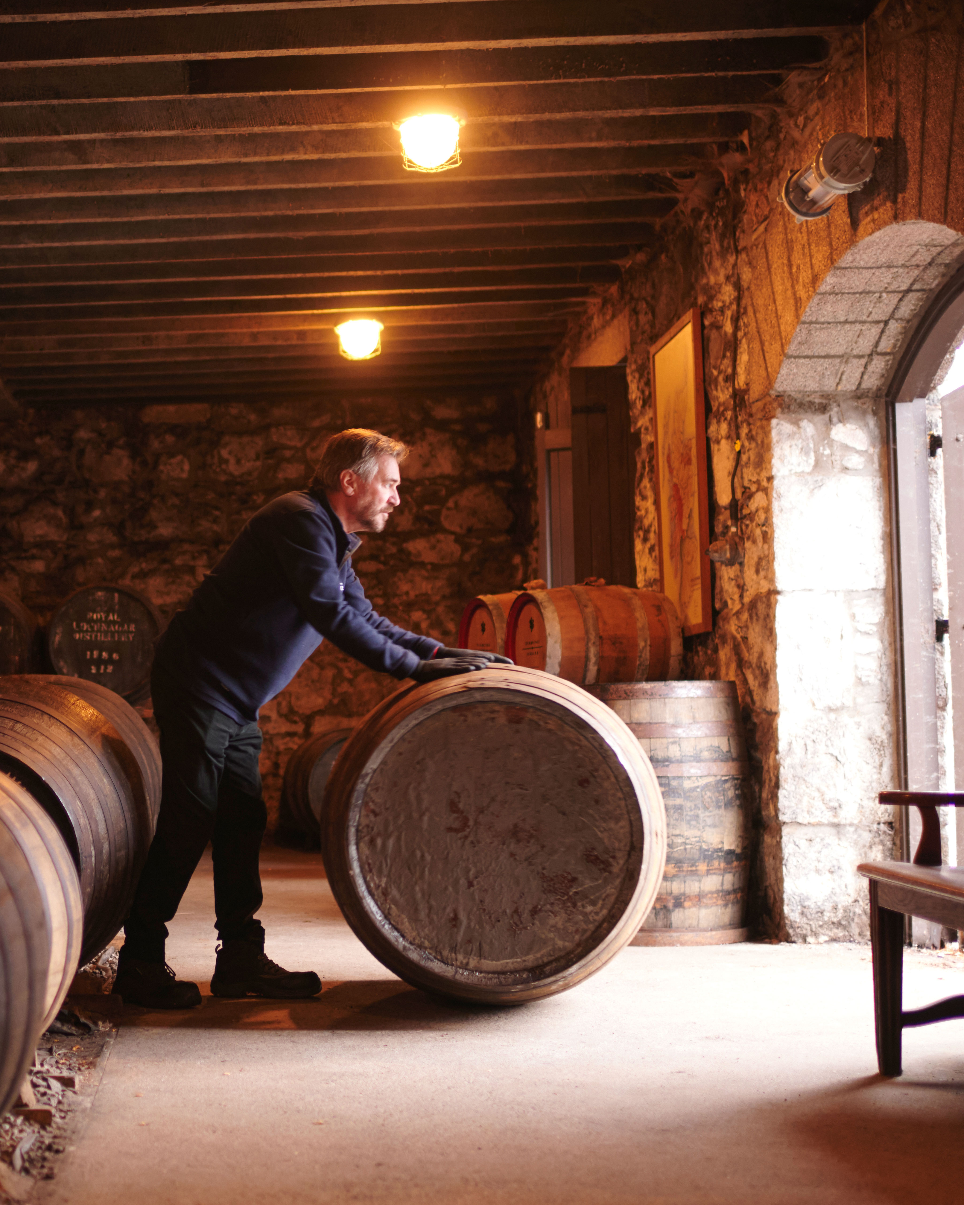 A man rolls a wooden barrel across the floor of a stone-walled cellar with several other barrels in the background.