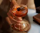 A Manhattan cocktail sits on a glass table, with a skewed maraschino cherry sitting over the rim of the coupe glass. A person holds the coupe by the stem.