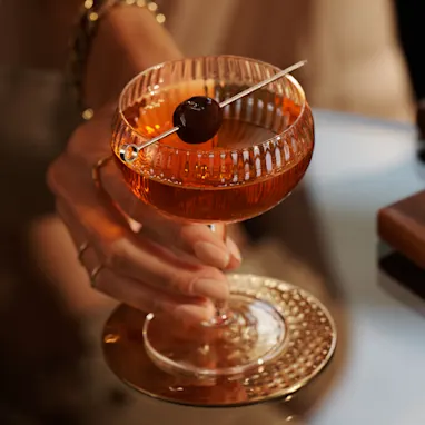 A Manhattan cocktail sits on a glass table, with a skewed maraschino cherry sitting over the rim of the coupe glass. A person holds the coupe by the stem.